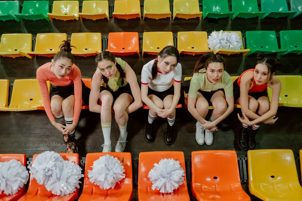 Group of asian cheerleaders sitting on colorful bleachers wearing sportswear with pompoms visible.