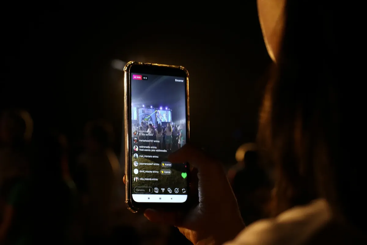 A woman live streams a concert using her smartphone at night. Vibrant stage in view.